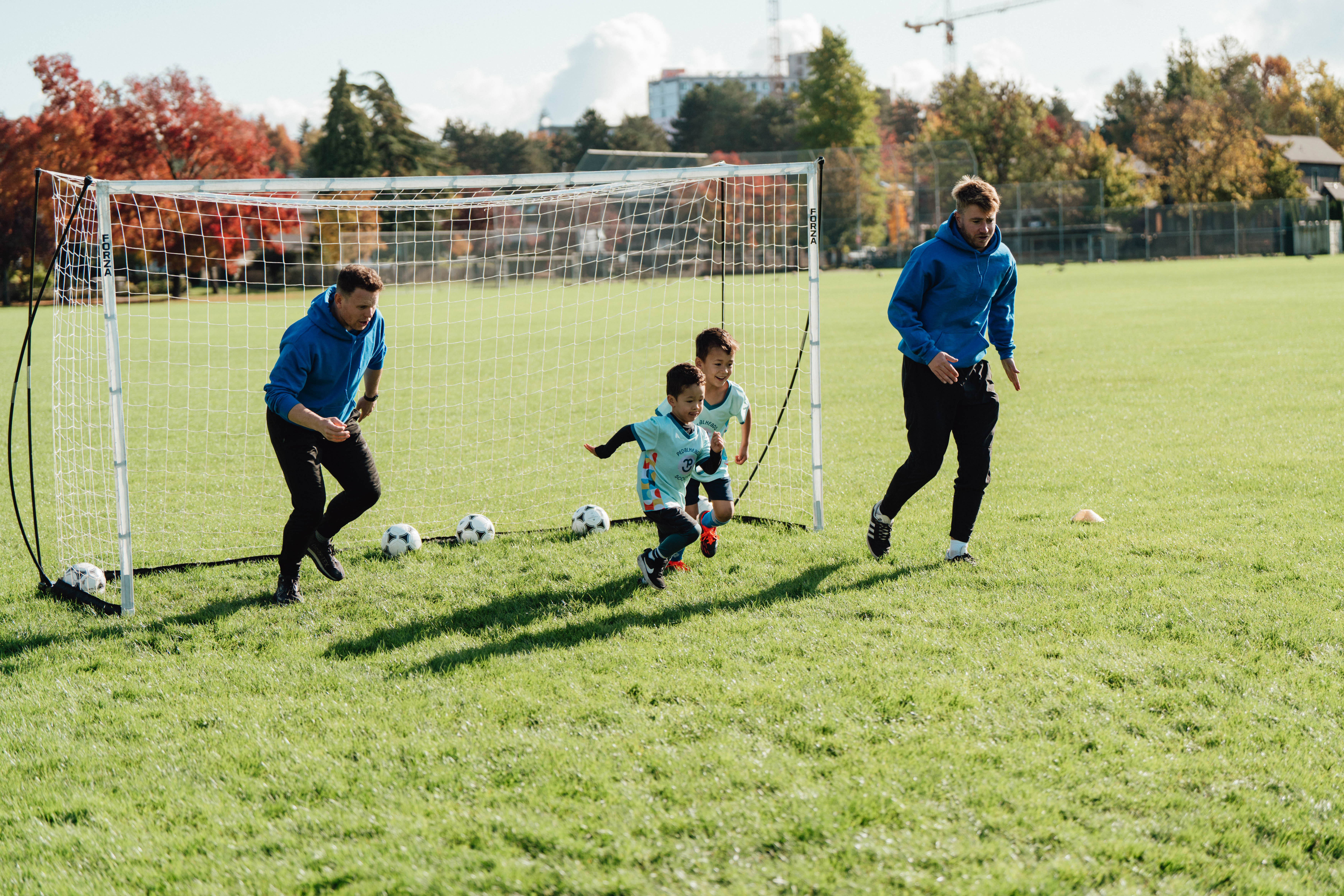 Des enfants courent avec des ballons de soccer loin d'un but lors d'un cours en plein air organisé par Pedalheads, sous la supervision d'entraîneurs.