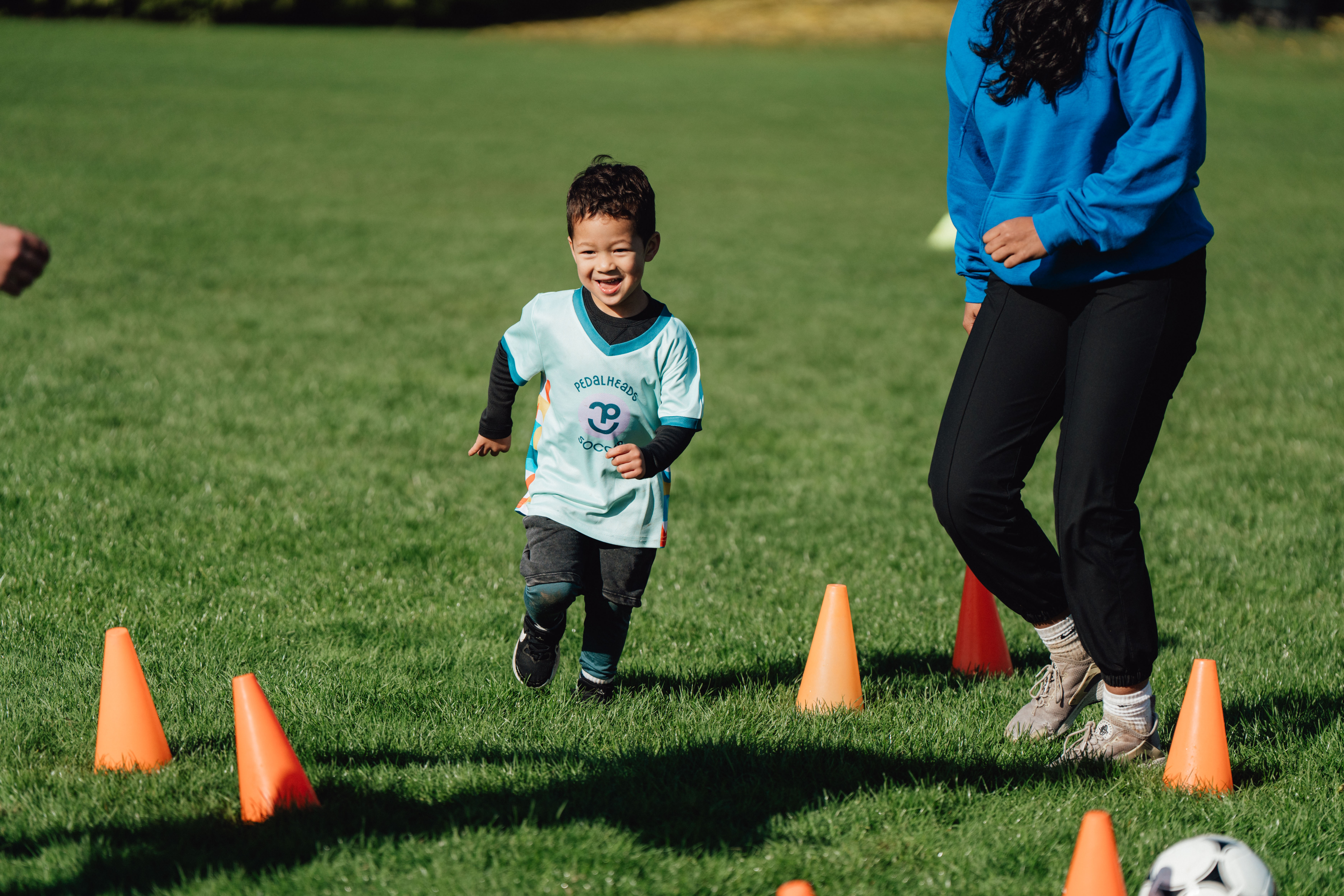 Un jeune enfant court entre des cônes orange lors d'une séance d'entraînement de soccer organisée par Pedalheads, sous la supervision d'une entraîneuse.
