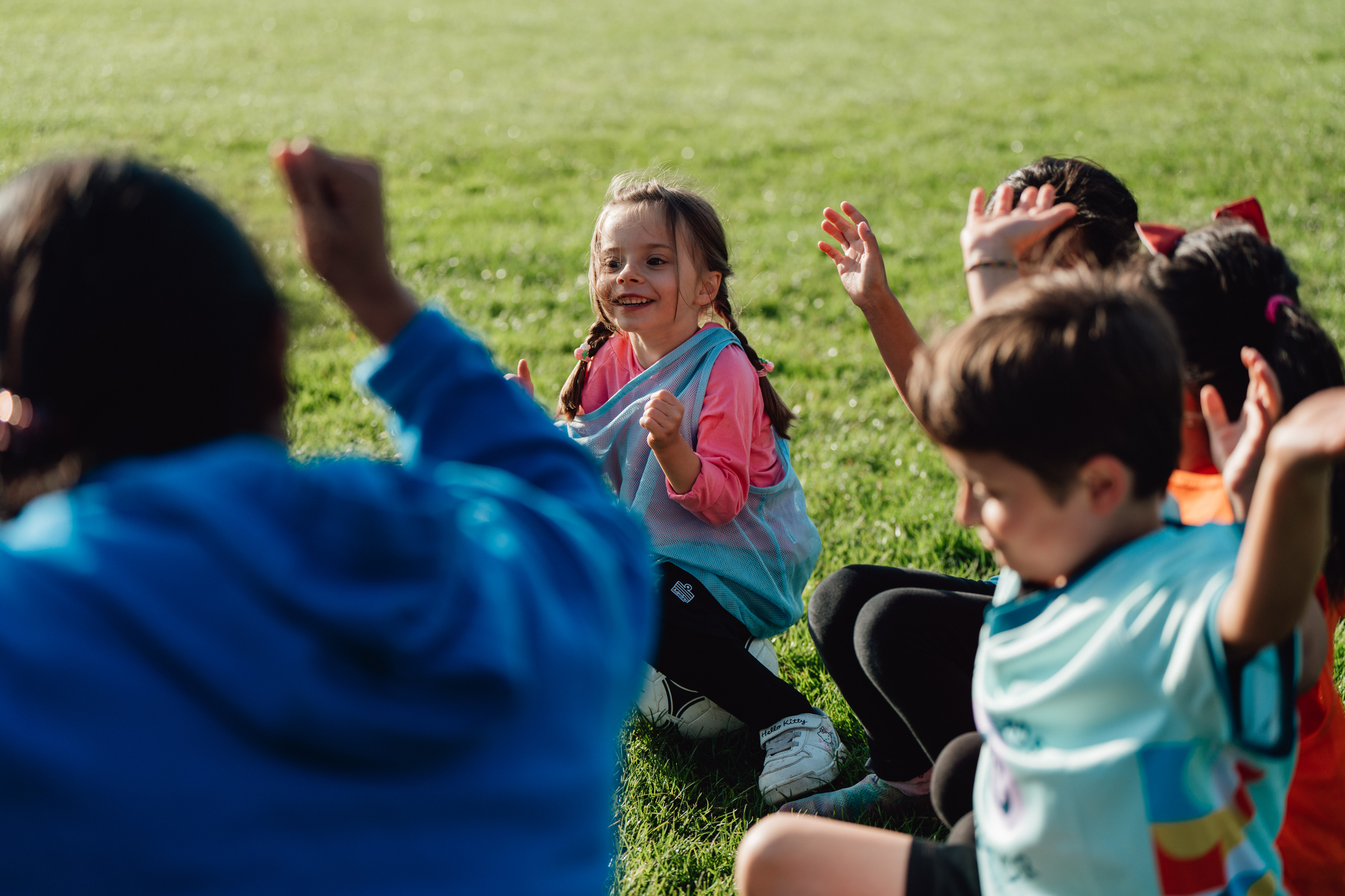 Des jeunes enfants participant à un cours de soccer organisé par Pedalheads, assis en cercle avec un entraîneur, levant la main et souriant pendant une activité de groupe.