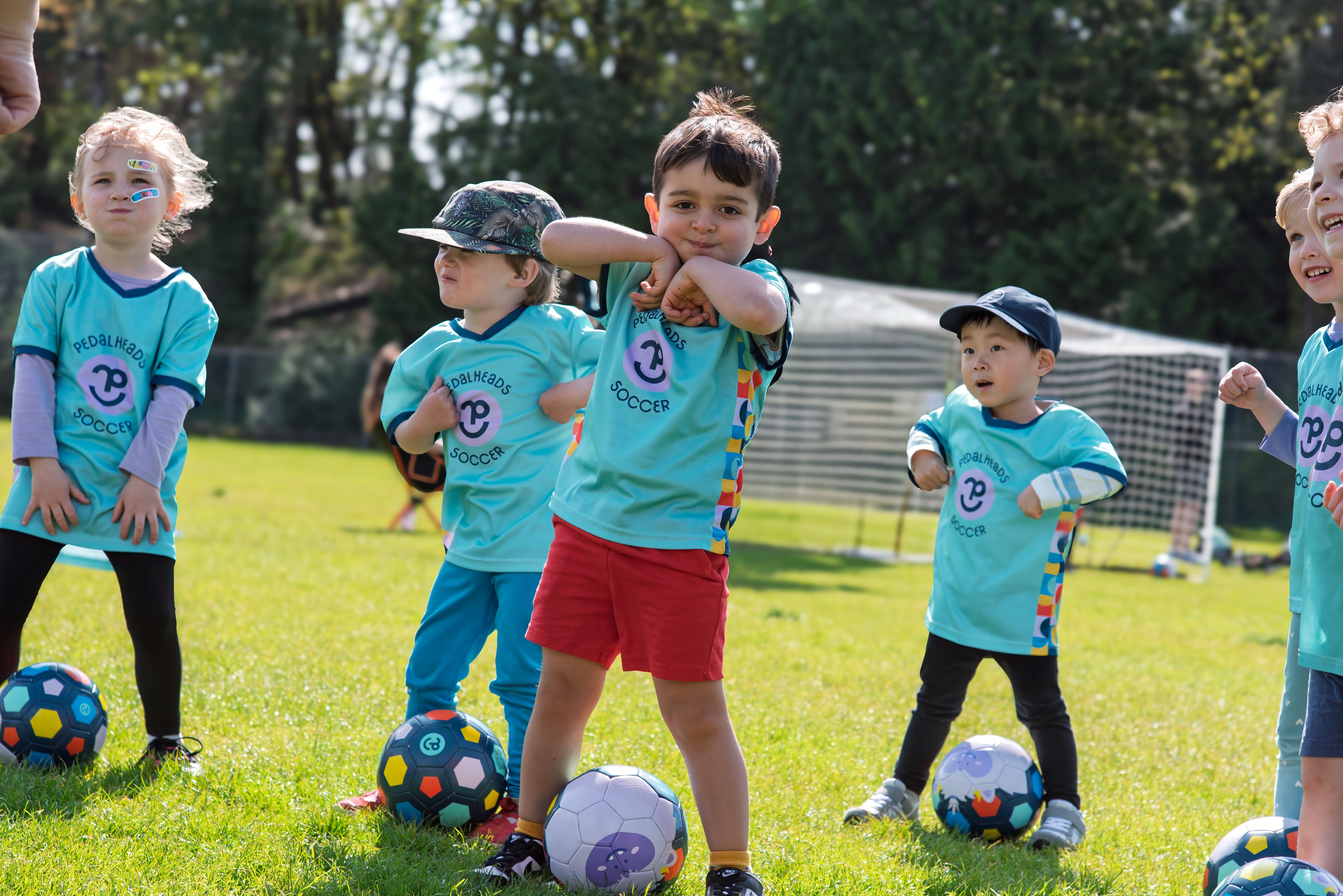 De jeunes enfants portant des maillots de soccer « Pedalheads », debout avec des ballons de soccer, participant à un exercice de motricité dirigé par un entraîneur.