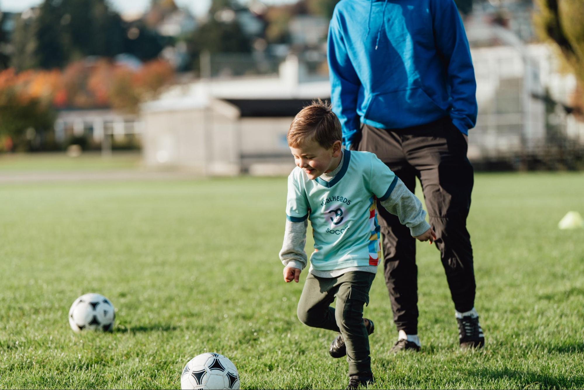 Un jeune enfant court vers un ballon sur un terrain de soccer pendant qu’un entraîneur marche juste derrière lui lors d’une séance d’entraînement.