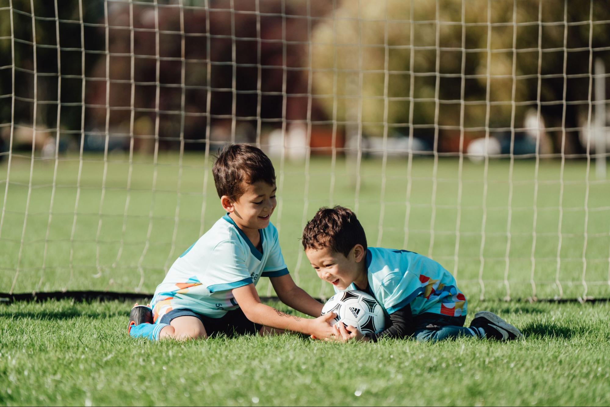 Deux jeunes enfants en tenue de soccer sourient et jouent avec un ballon assis devant un but sur un terrain ensoleillé.